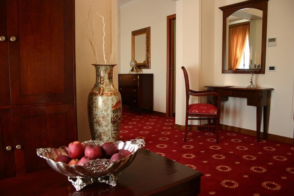 A room with a wooden door, a large vase with dried branches, a mirror, a red patterned carpet, and a table with a fruit bowl.
