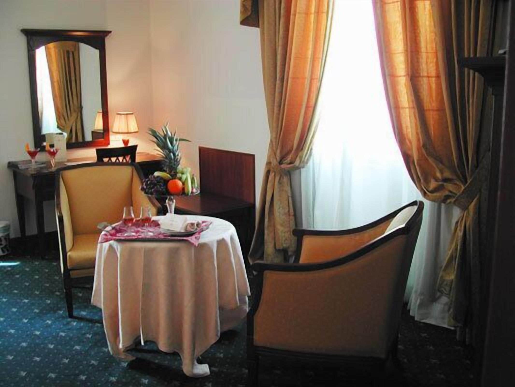 Habitación de hotel con una mesa de comedor para dos, una silla, una mesita con un frutero y una lámpara.