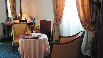 Habitación de hotel con una mesa de comedor para dos, una silla, una mesita con un frutero y una lámpara.