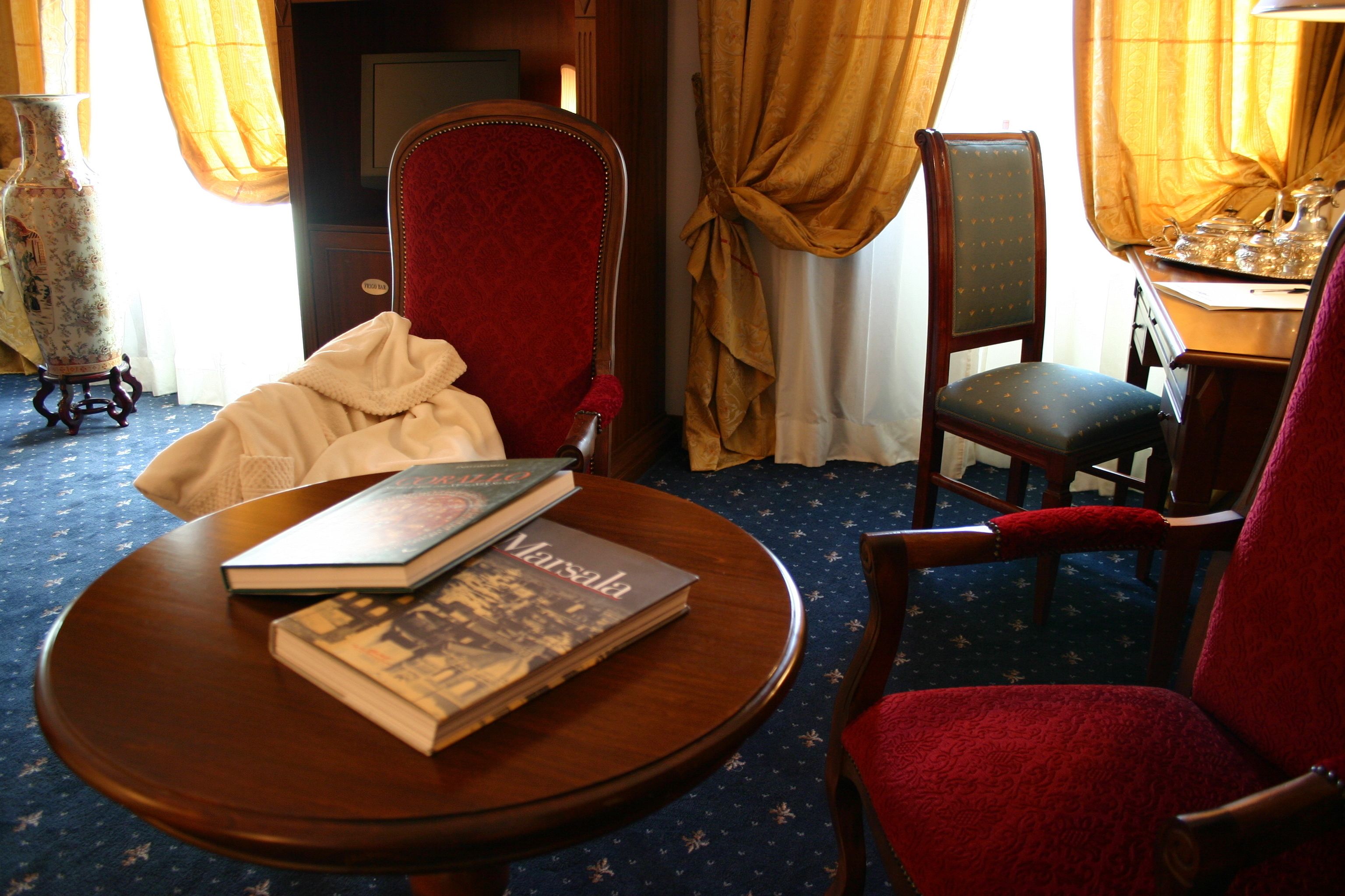 A room with a round wooden table, two red upholstered chairs, a bookshelf, and a large vase.