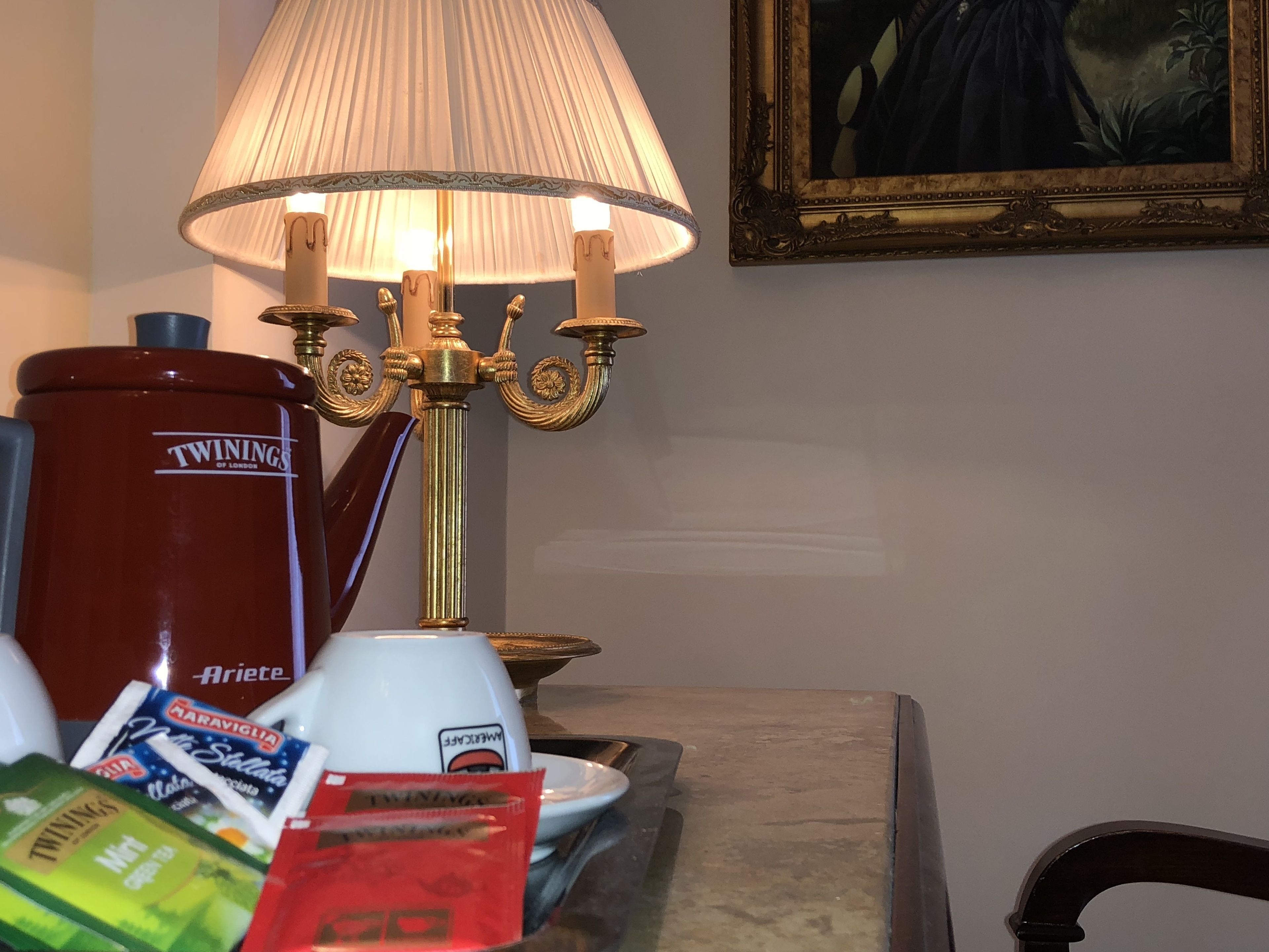 A table with a red coffee maker, a white coffee pot, a lamp, and some food items.