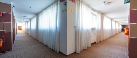 Un pasillo de hotel con piso alfombrado, cortinas blancas y señalización que indica un piano y una salida.