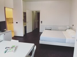 A hotel room with a bed, bedside table, and a desk with a chair. There is a bathroom with a sink and mirror.