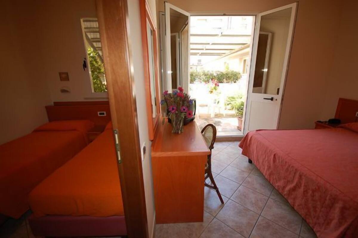 A room with two beds, a desk, and a chair. There is a door leading to an outdoor area.