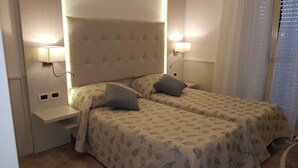 A room with two beds, each with a headboard and bedside lamps. There is a small shelf unit under the headboard and a window with blinds. - Hotel Bella Riviera (Viareggio)