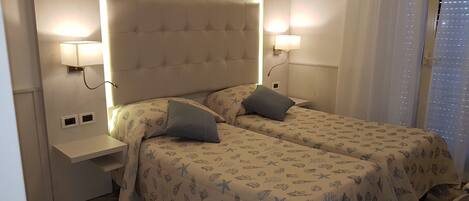 A room with two beds, each with a headboard and bedside lamps. There is a small shelf unit under the headboard and a window with blinds.
