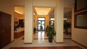 A hotel lobby with a reception desk, a large potted plant, and a tiled floor. - Hotel Bella Riviera (Viareggio)