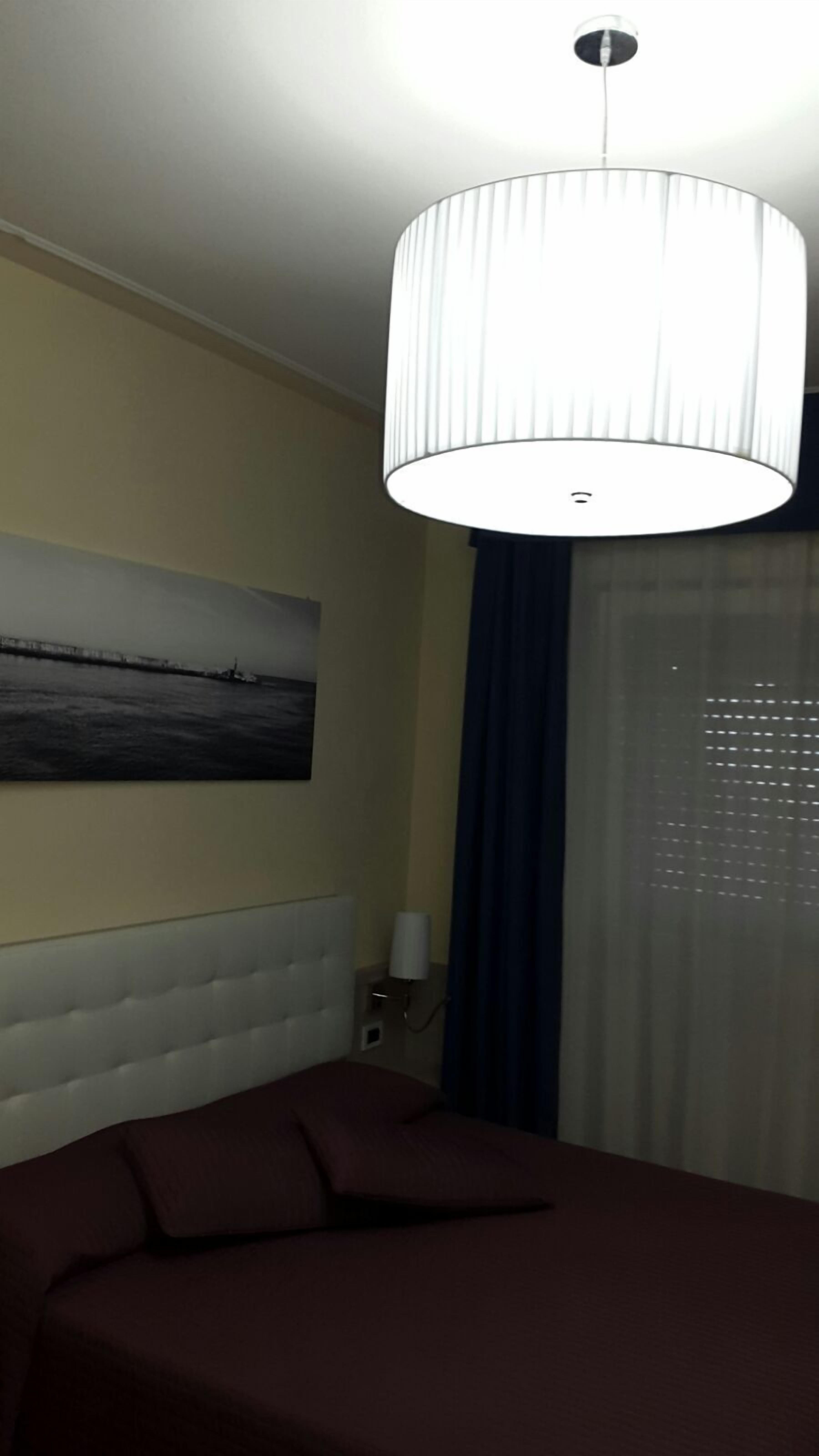 A hotel room with a bed, a headboard, a wall-mounted lamp, a painting, and a window with curtains.