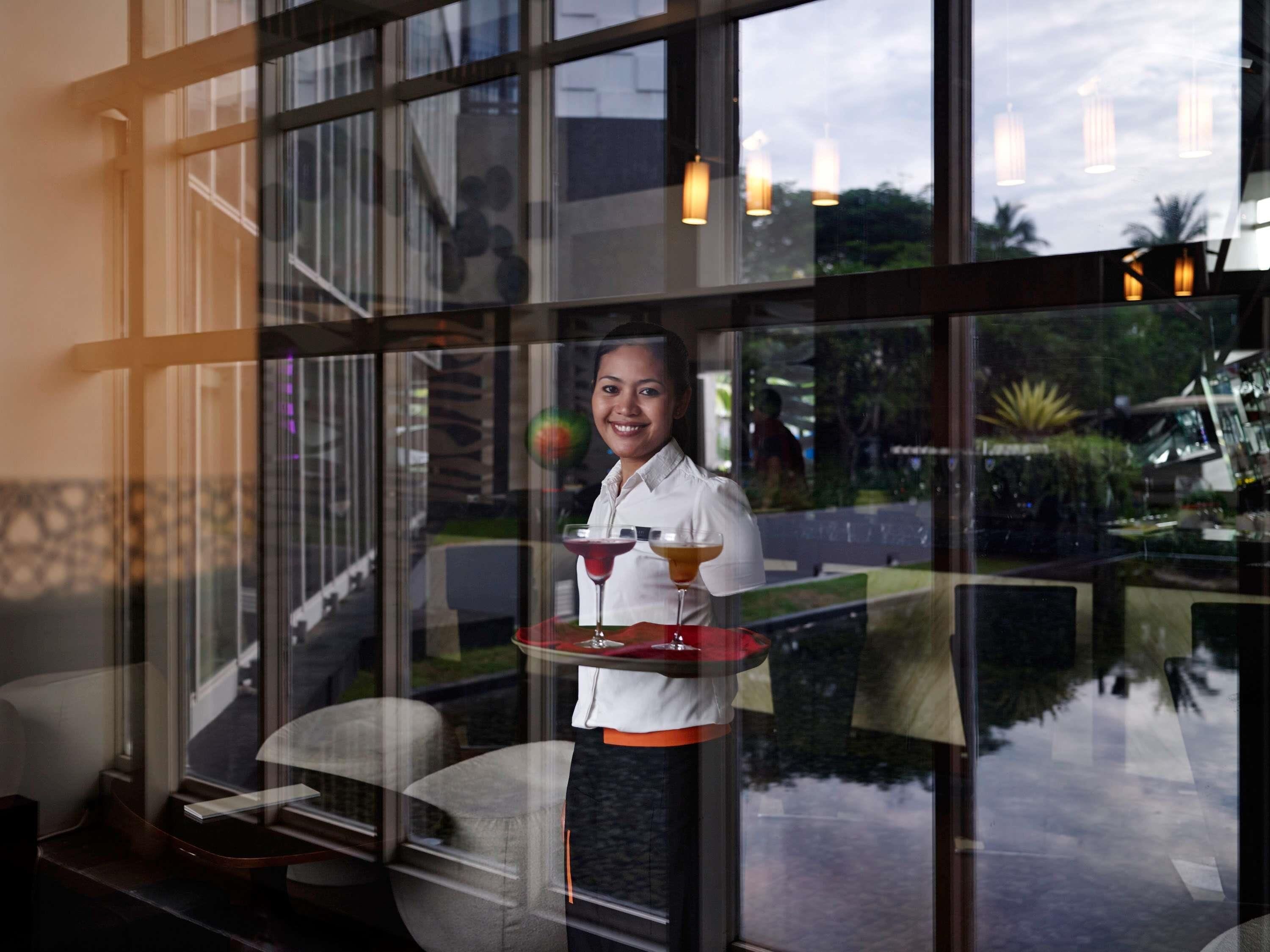 A person holding a tray with two drinks, standing in front of a window with a view of outdoor seating and greenery.