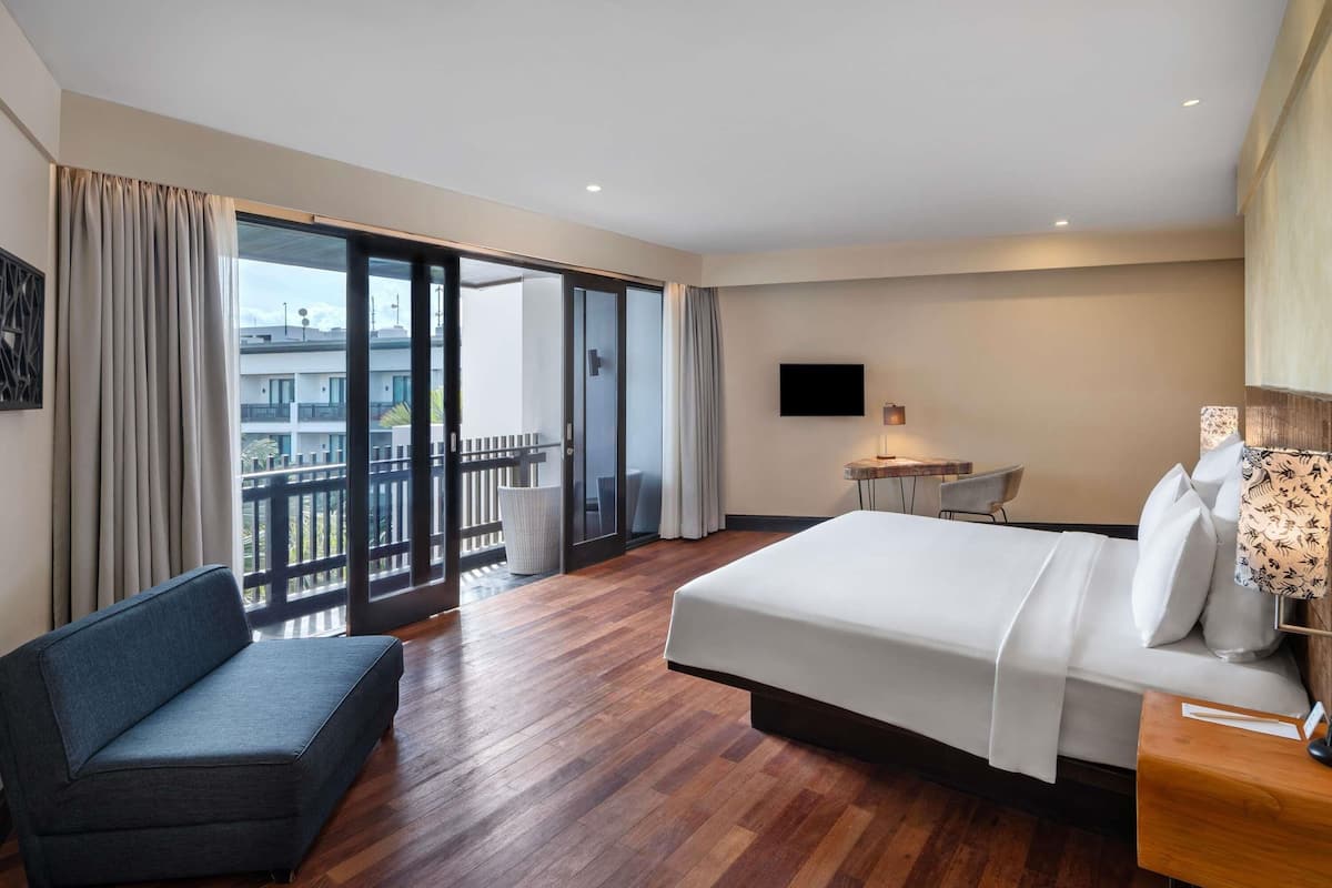A hotel room with a large bed, a sofa, a desk, and a balcony with a view of buildings.