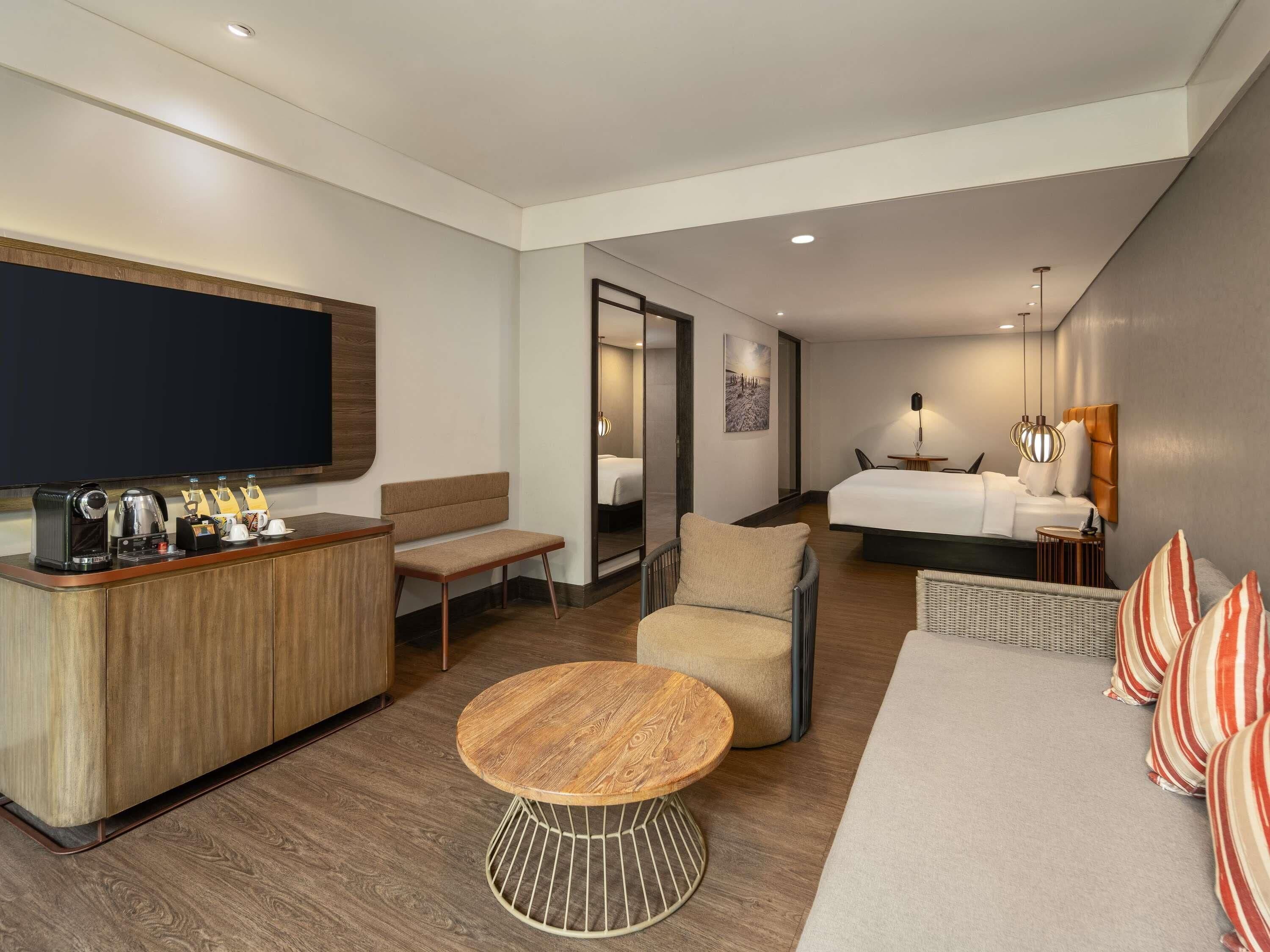 A modern hotel room with a wooden desk, a round coffee table, a sofa, a bed, and a television.