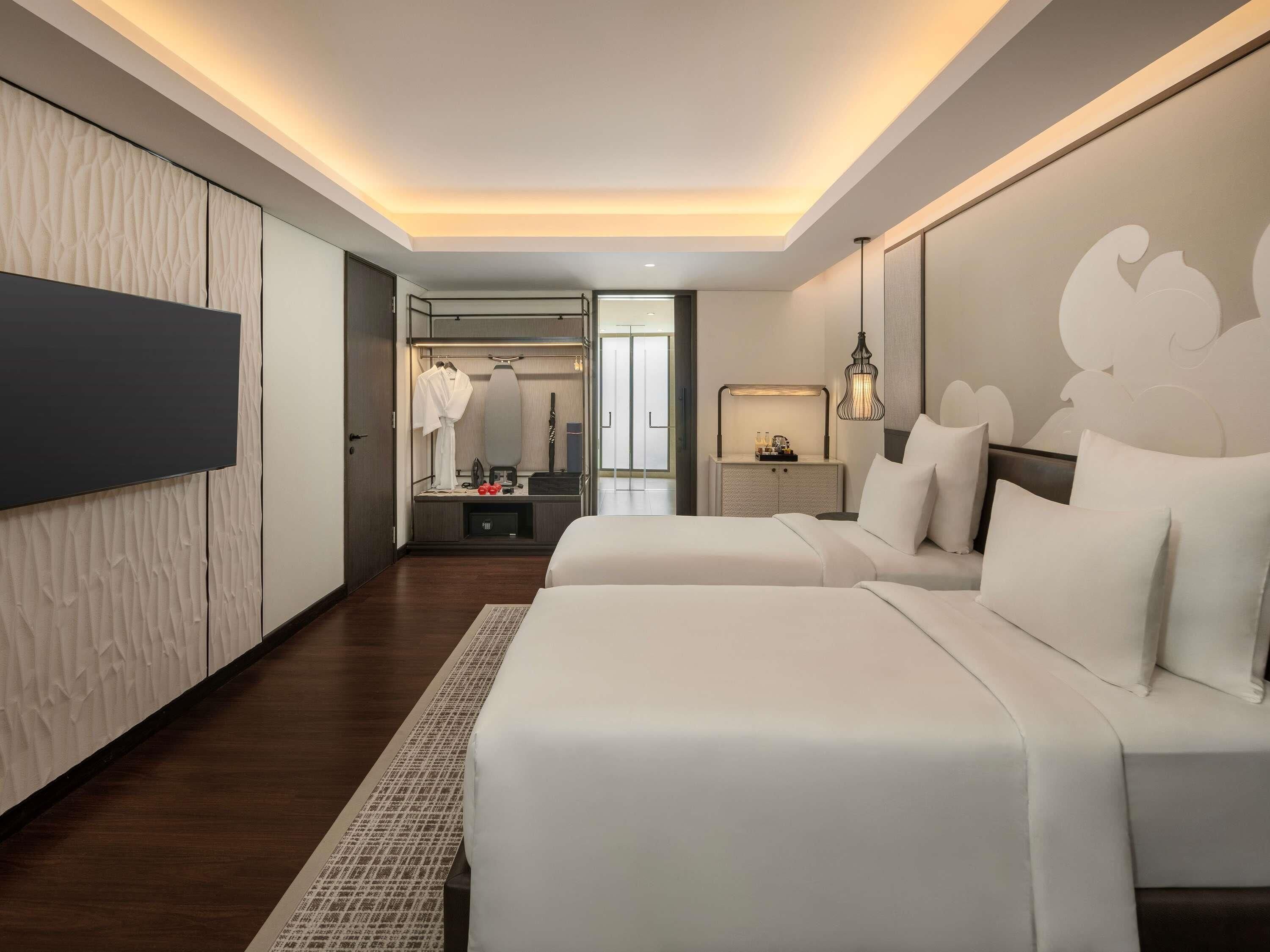 Quarto de hotel moderno com duas camas, um guarda-roupa, uma toucador com espelho e uma mesa de cabeceira com abajur.