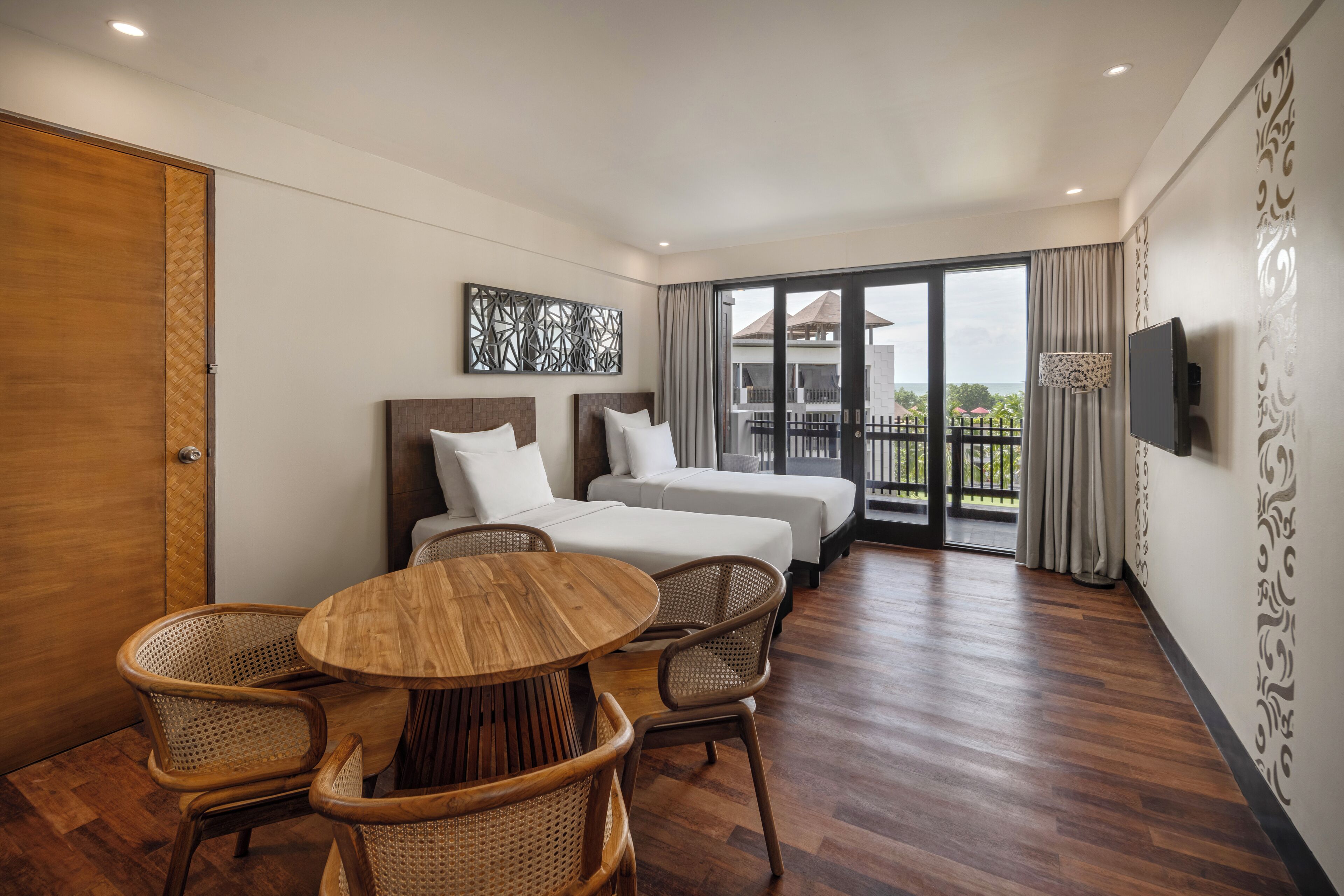A hotel room with a wooden floor, a round wooden table with chairs, a bed with white bedding, a TV mounted on the wall, and a balcony with a view.