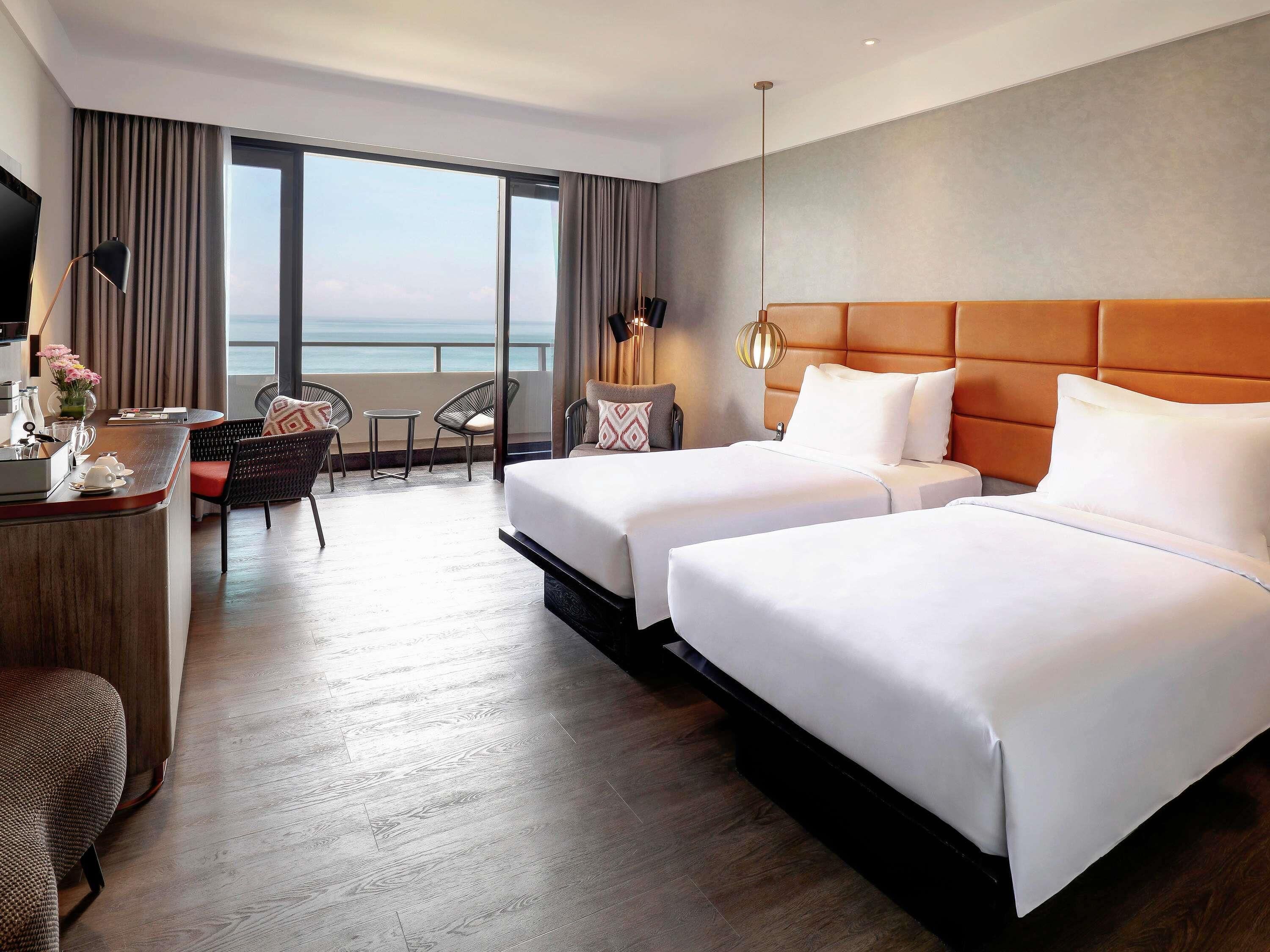 A hotel room with two beds, a desk, a chair, and a view of the sea.