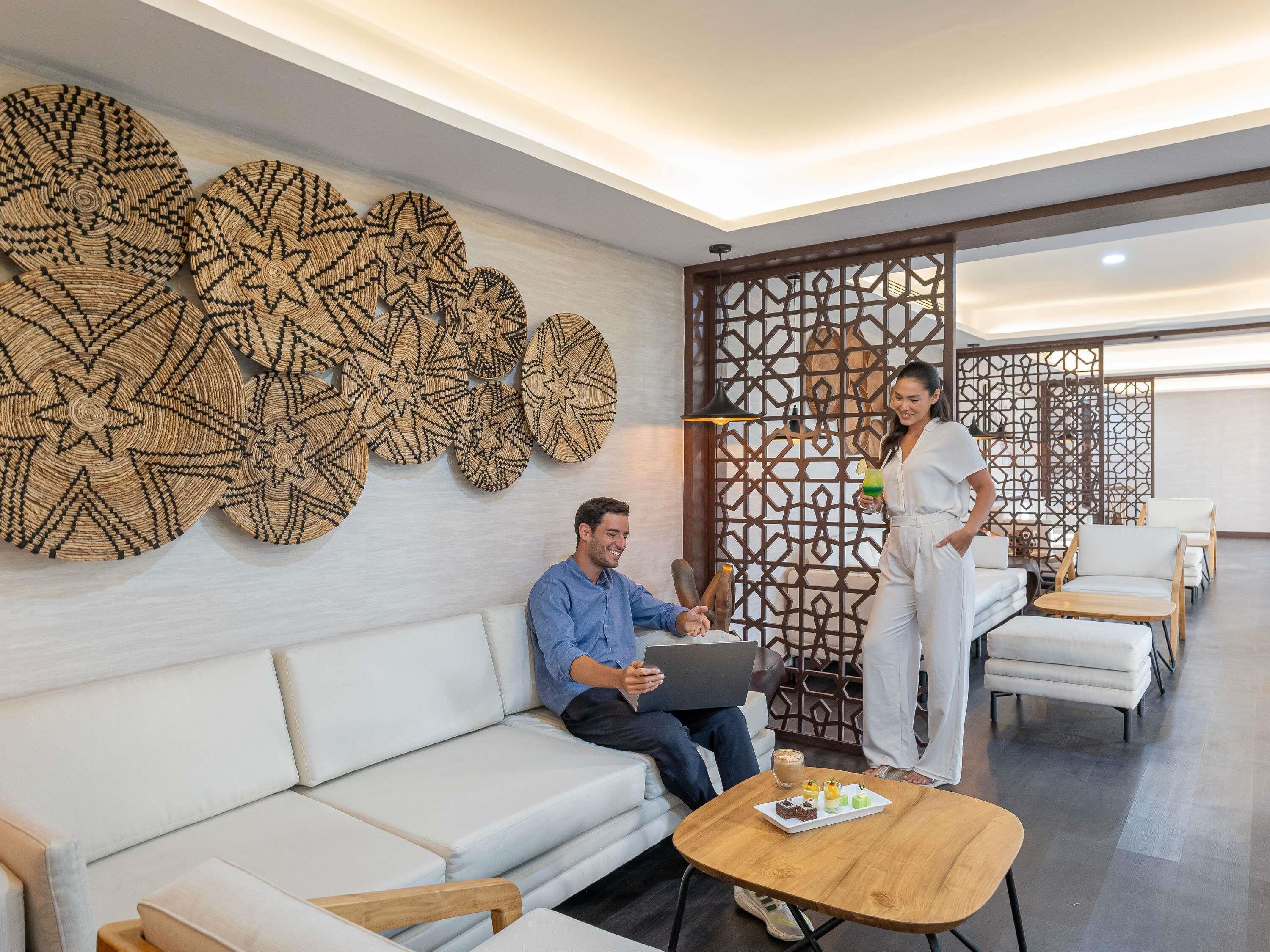 Un moderno hall de hotel con un sofá blanco, una mesa de centro de madera con postres y una pared decorativa con motivos tejidos.