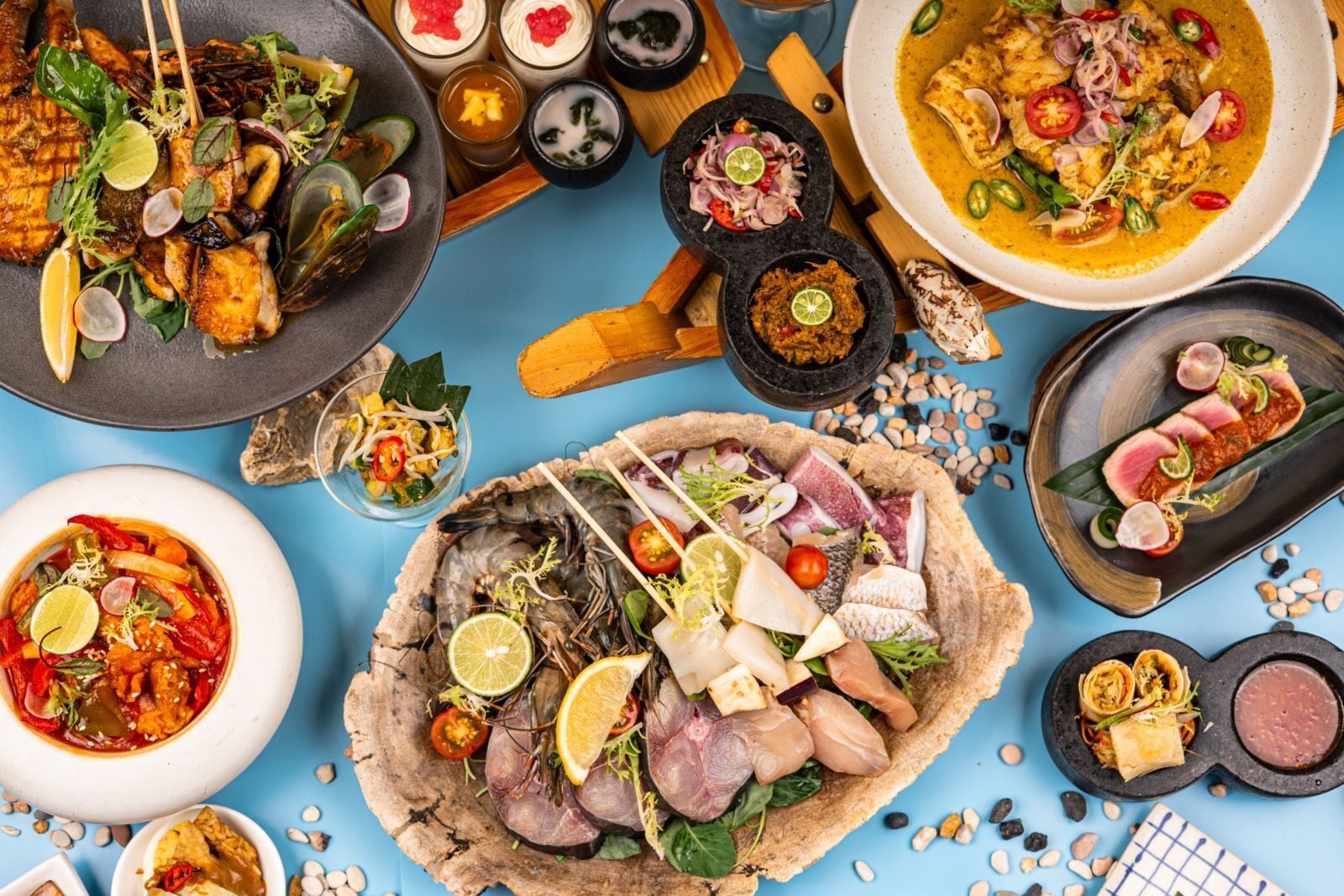 Variedad de platos, incluyendo pescado a la parrilla, mariscos y un bol de sopa, sobre una superficie azul.