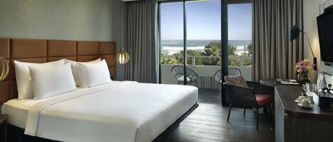 A hotel room with a large bed, a desk with a chair, a TV, and a balcony with a view of the ocean.