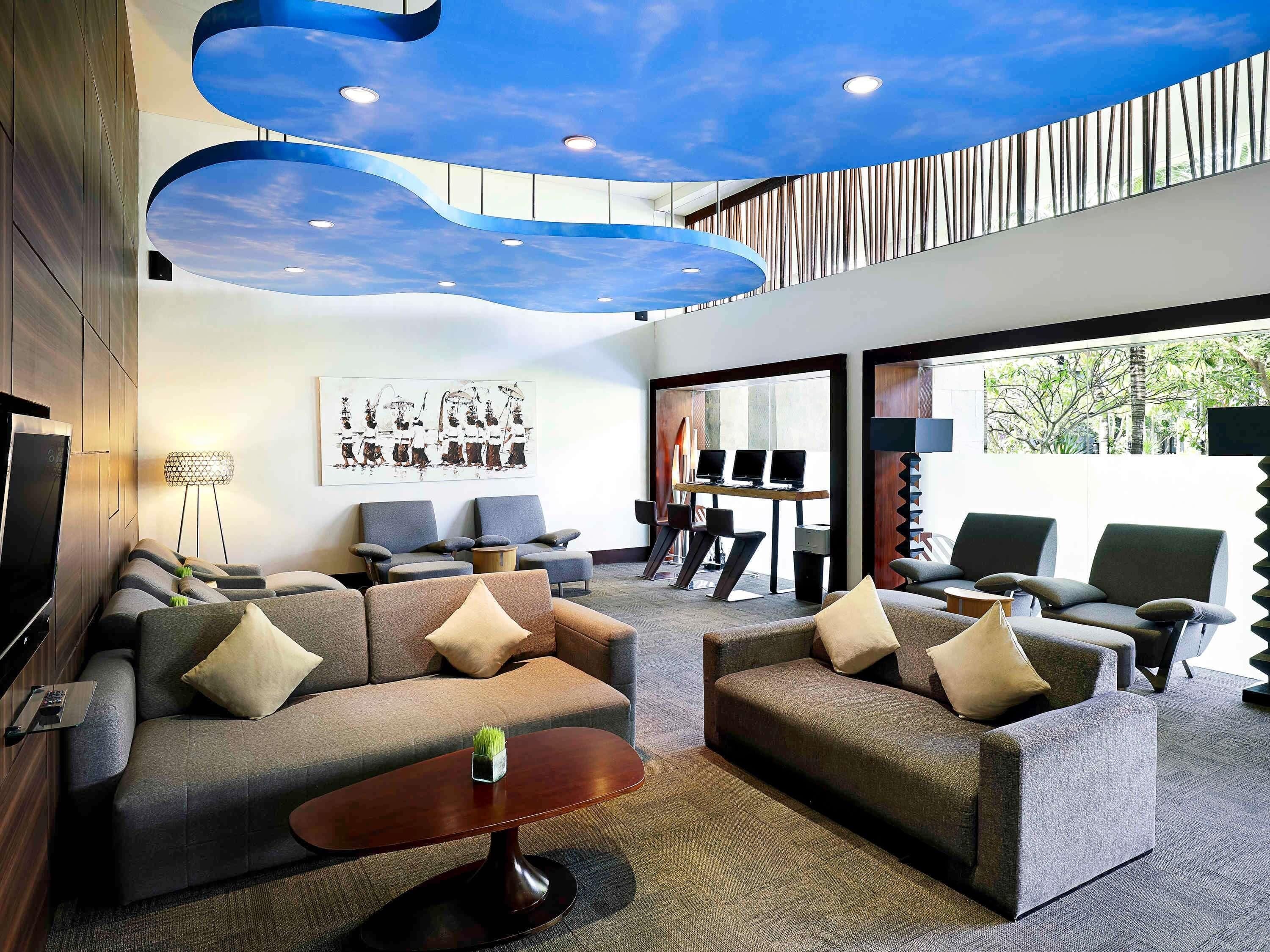 A modern interior with sofas, a coffee table, a desk with computers, and a ceiling with a blue sky design.