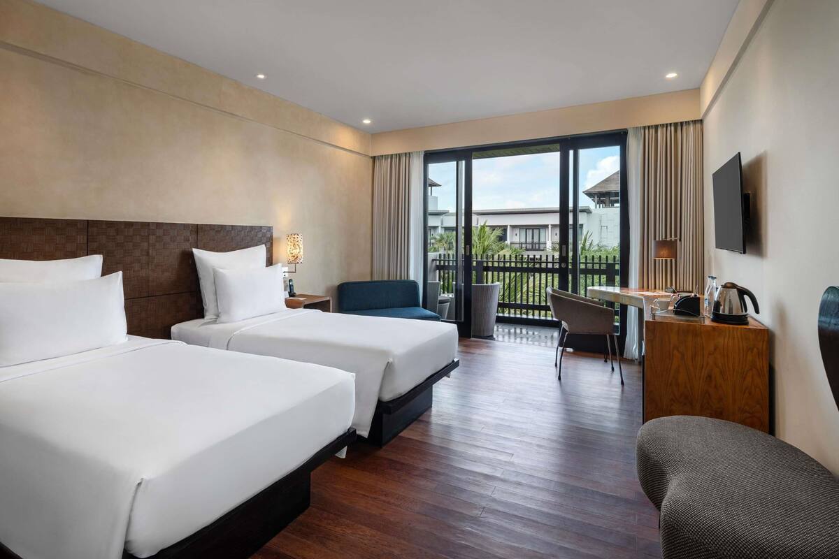 A hotel room with two beds, a desk, a chair, a TV, and a balcony with a view.