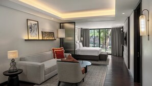Quarto de hotel moderno com sofá, poltrona, mesa de cabeceira, luminária e uma cama com vista para o exterior.