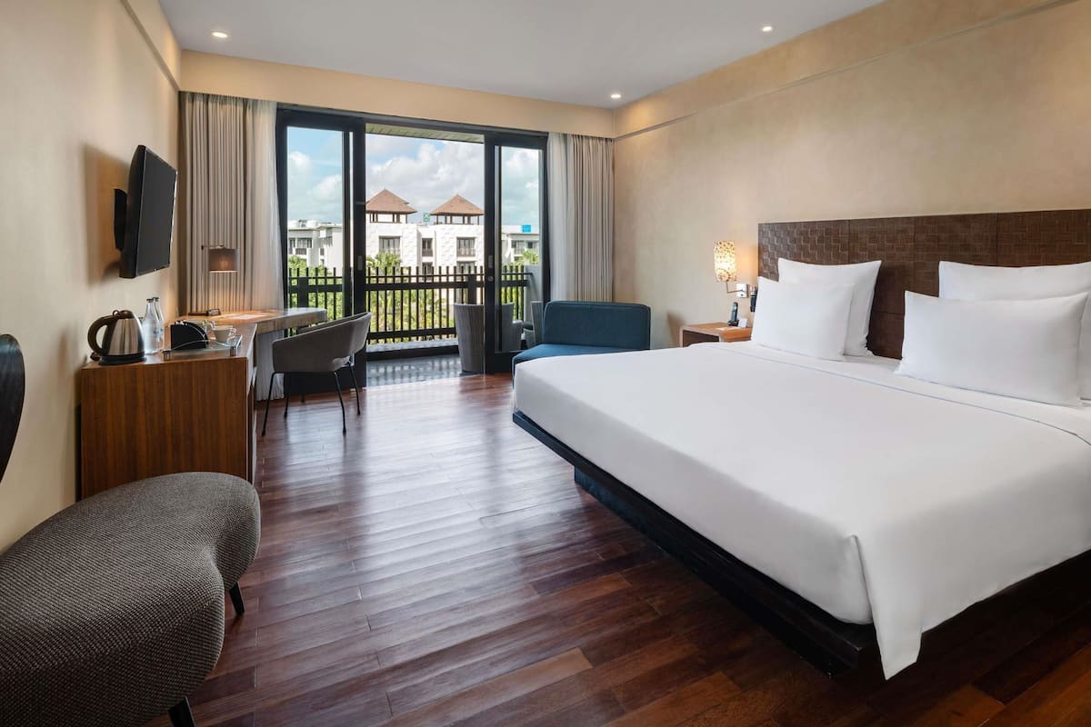A hotel room with a large bed, a desk with a chair, a television, and a balcony with a view of buildings.