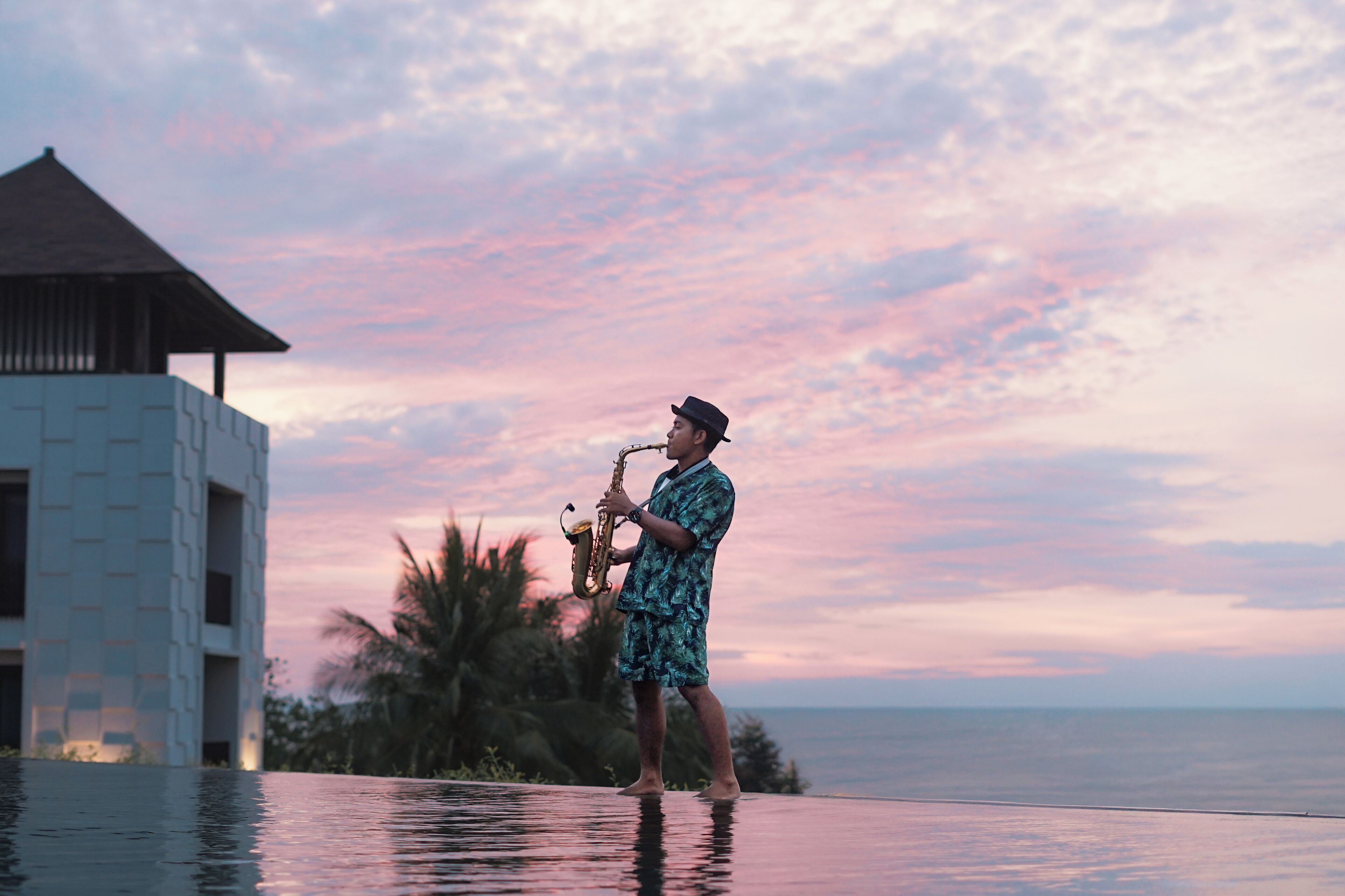 Una persona suona il sassofono a bordo piscina con il sole al tramonto sullo sfondo.