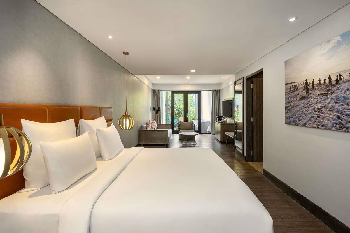 A modern hotel room with a large bed, a sofa, a dining area, and a view of the outdoors.