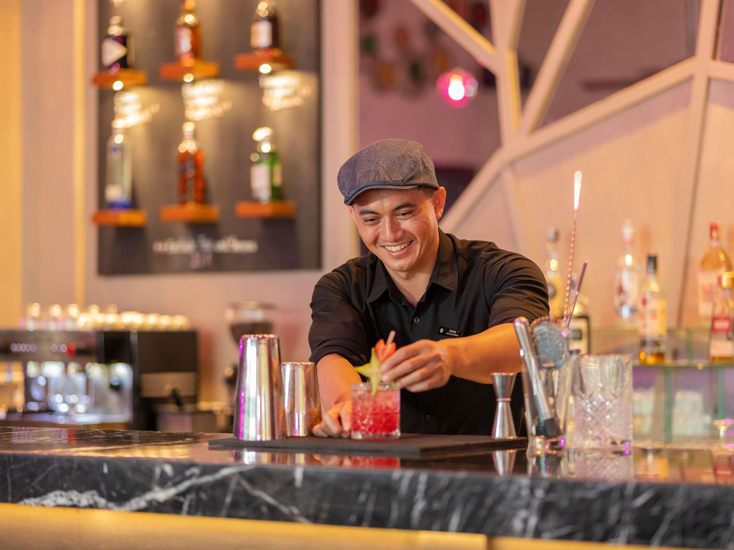 Un bar con un cantinero preparando una bebida, instrumentos de bar y botellas al fondo.