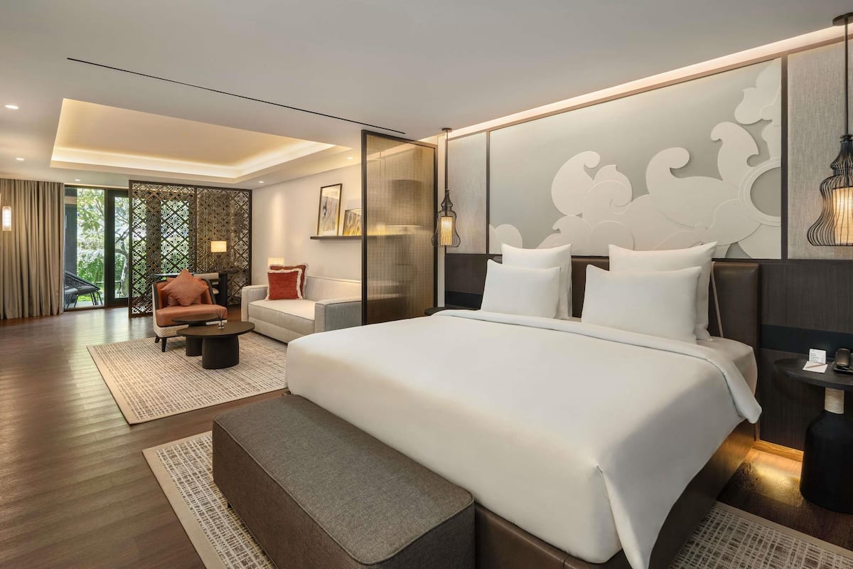 A modern hotel room with a large bed, a sitting area with a sofa and armchair, a small table, and a decorative wall panel.
