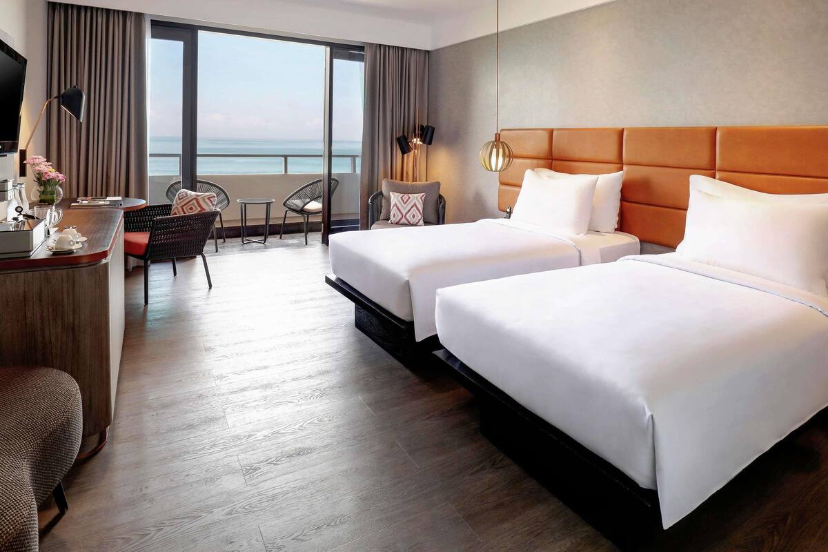 A hotel room with two beds, a desk, a chair, and a view of the sea.