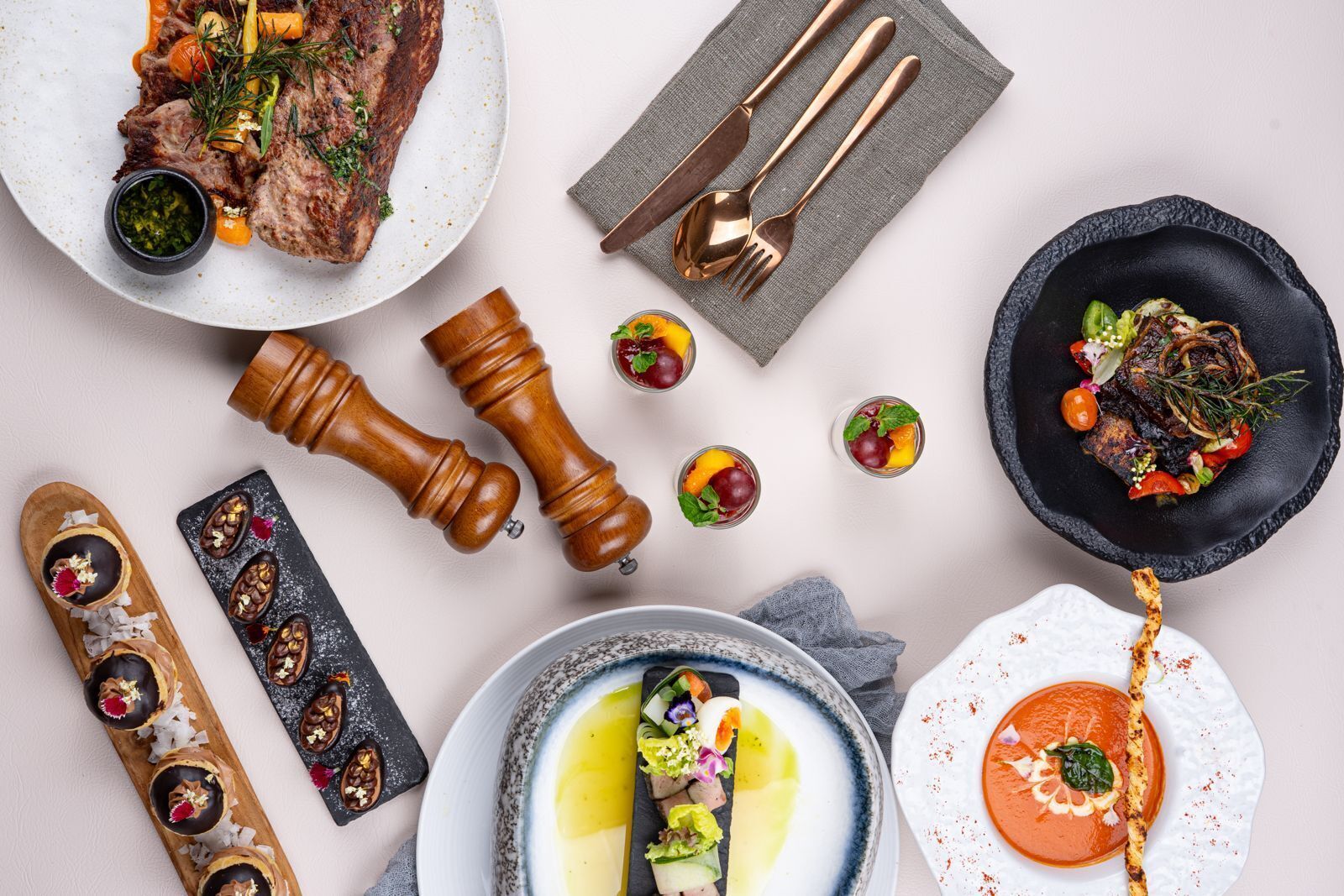A variety of dishes including a steak, a bowl of soup, and a plate with a tomato-based dish, along with some condiments and a napkin.