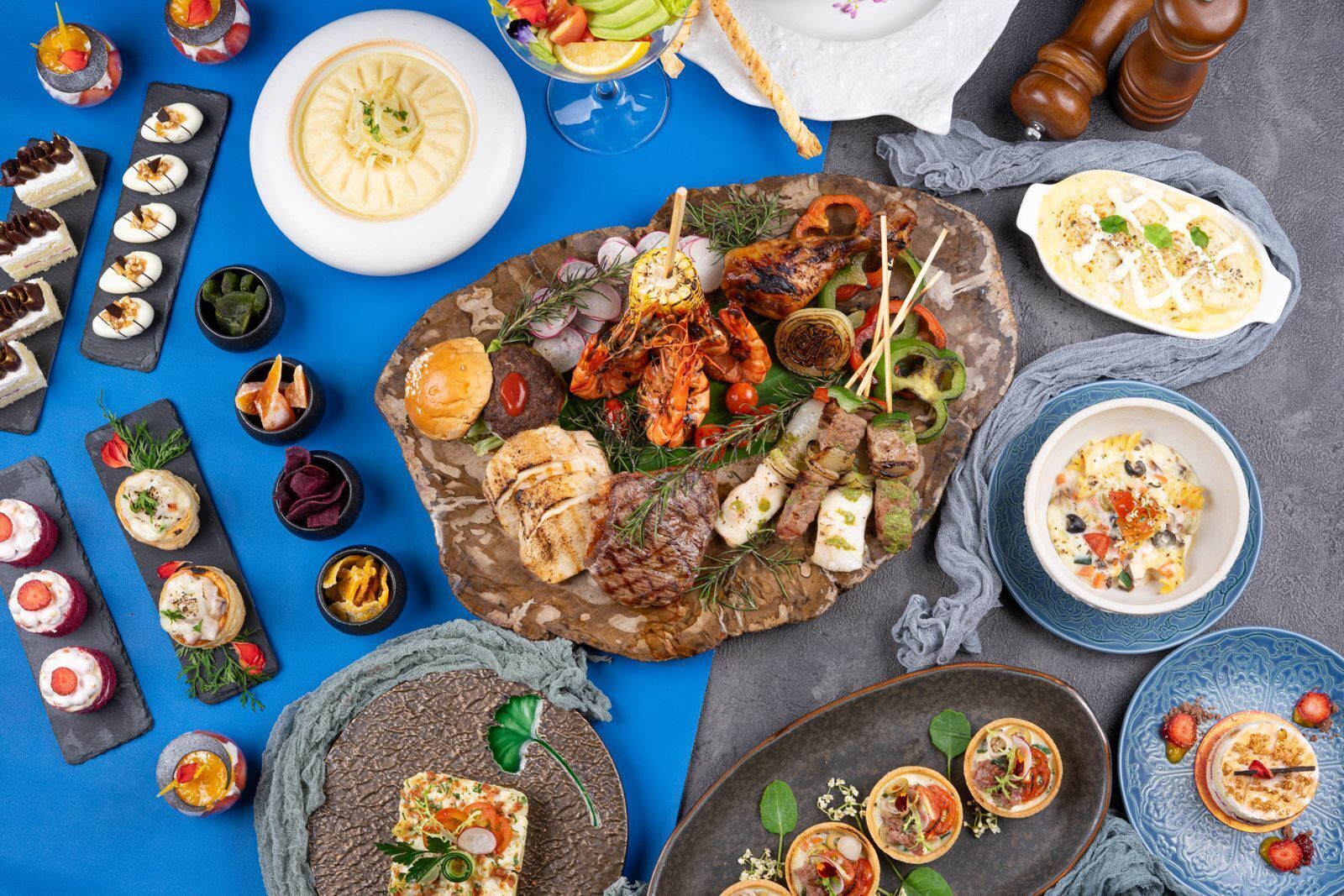 Variedad de alimentos, incluyendo carnes a la parrilla, verduras y postres, sobre una superficie azul.