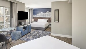 Habitación de hotel con cama, televisor, escritorio, silla, una mesita y vistas a la ciudad.
