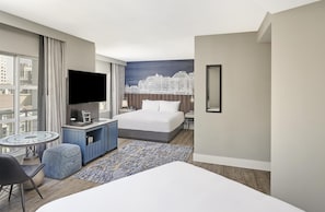 Habitación de hotel con cama, televisor, escritorio, silla, una mesita y vistas a la ciudad.