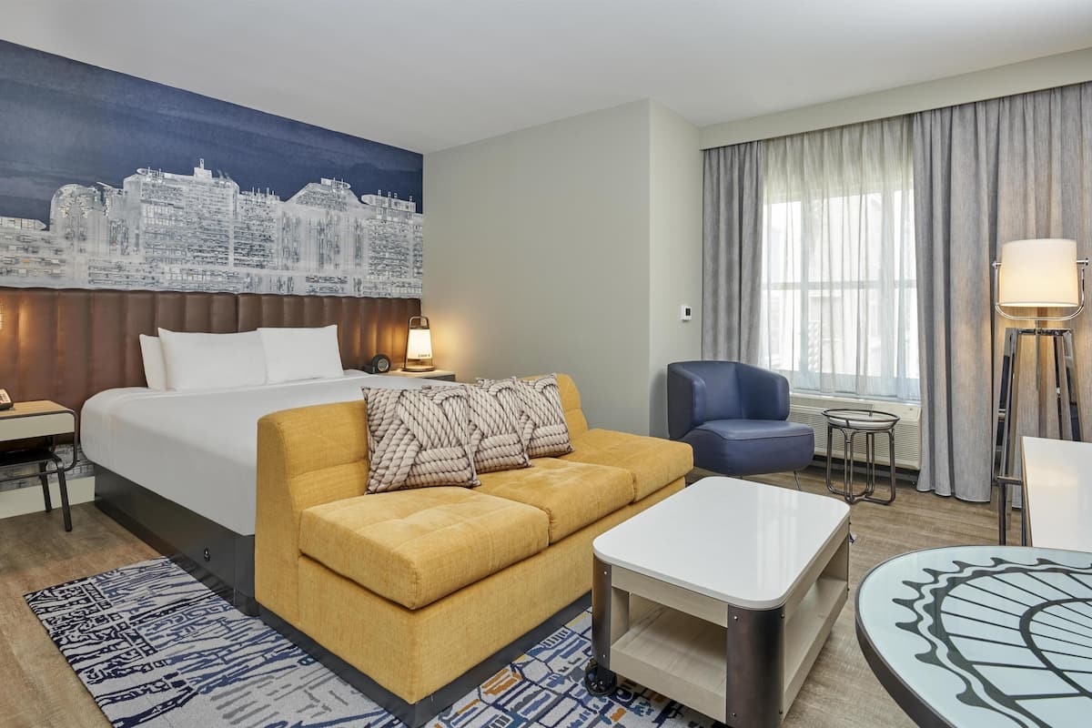A hotel room with a bed, a sofa, a coffee table, a desk, a chair, a lamp, and a cityscape mural on the wall.