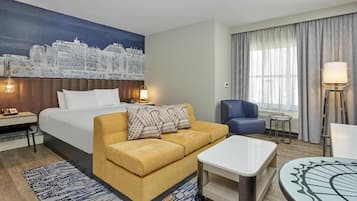 A hotel room with a bed, a sofa, a coffee table, a desk, a chair, a lamp, and a cityscape mural on the wall.