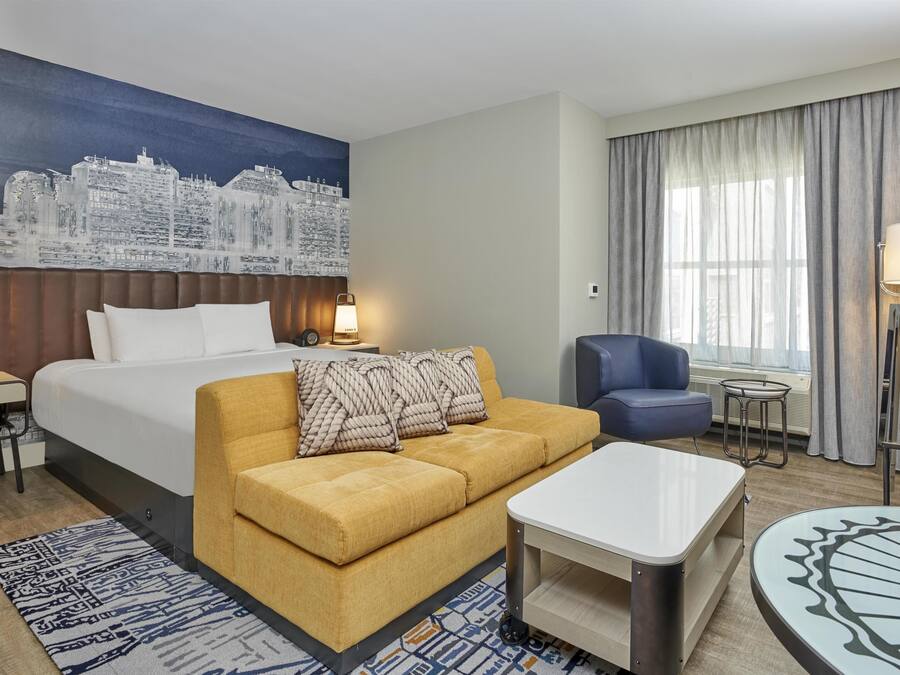 A hotel room with a bed, a sofa, a coffee table, a desk, a chair, a lamp, and a cityscape mural on the wall.
