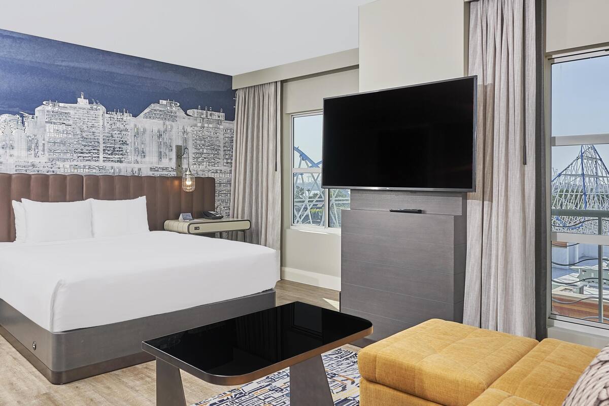 A hotel room with a bed, a desk, a TV, and a large window with a cityscape view.