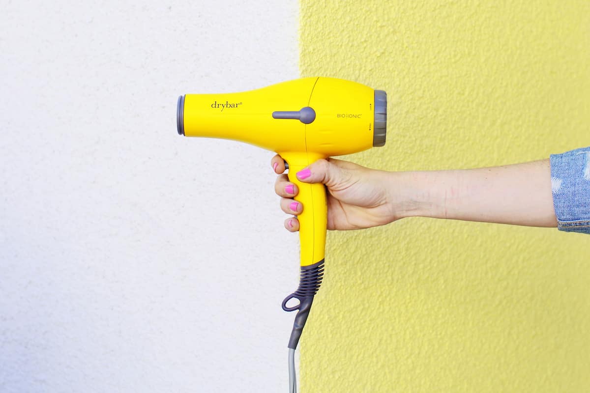 A hand holding a yellow hairdryer against a yellow wall.
