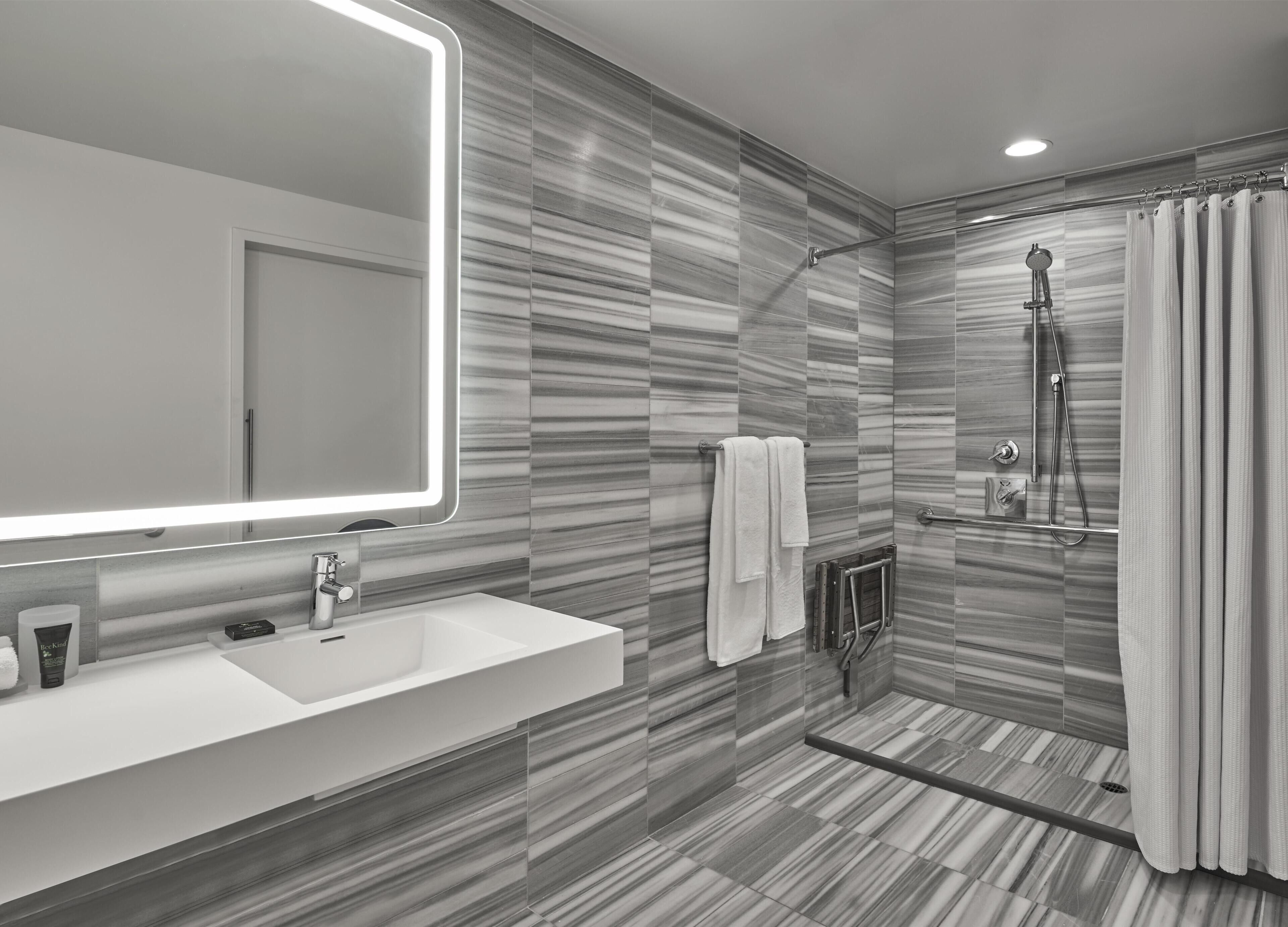 Une salle de bain moderne avec un grand miroir, un lavabo blanc, une douche équipée d’un pommeau de douche à main et un porte-serviettes.