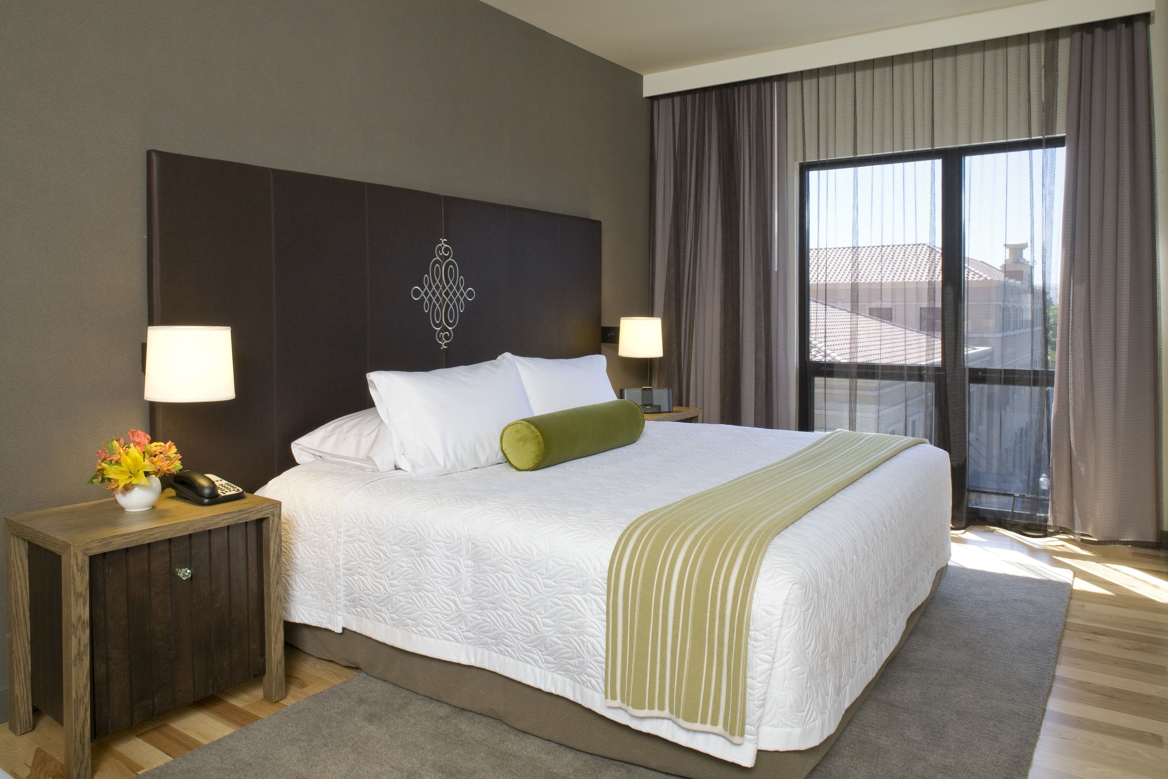 A hotel room with a large bed, two bedside tables, a nightstand, a decorative wall piece, and a large window with curtains.