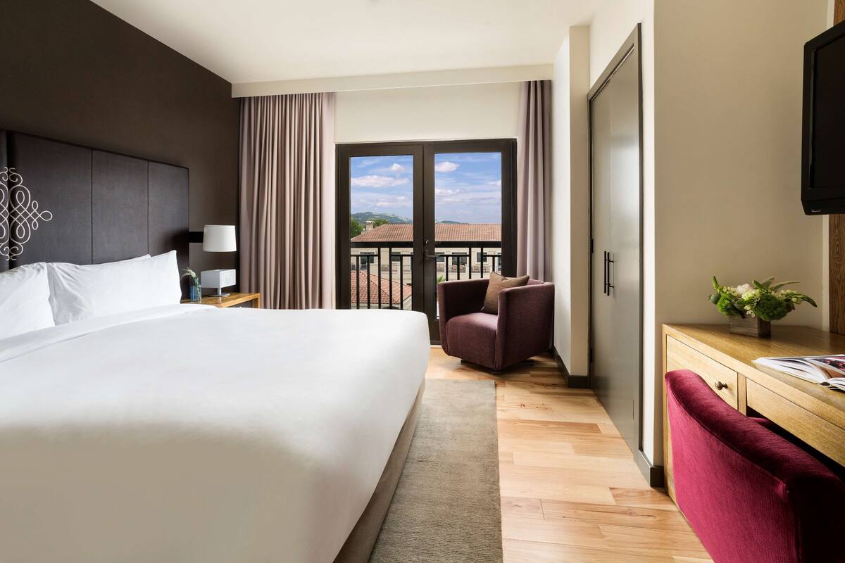 A hotel room with a large bed, a desk with a chair, a TV, and a balcony with curtains.