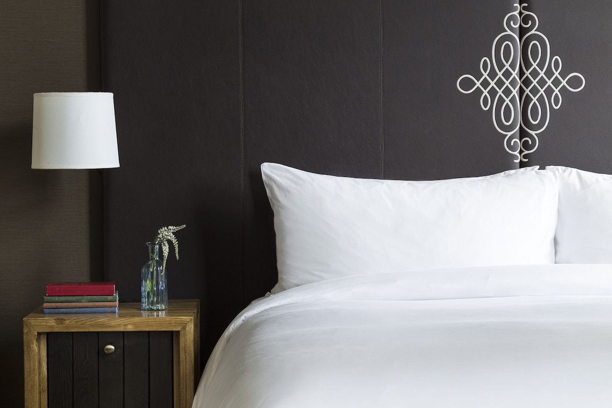 A neatly made bed with white linens, a dark headboard, and a bedside table with a lamp and books.