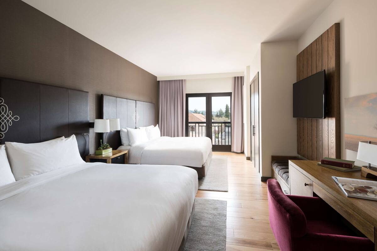 A hotel room with two beds, a desk, a TV, and a view of the outdoors.