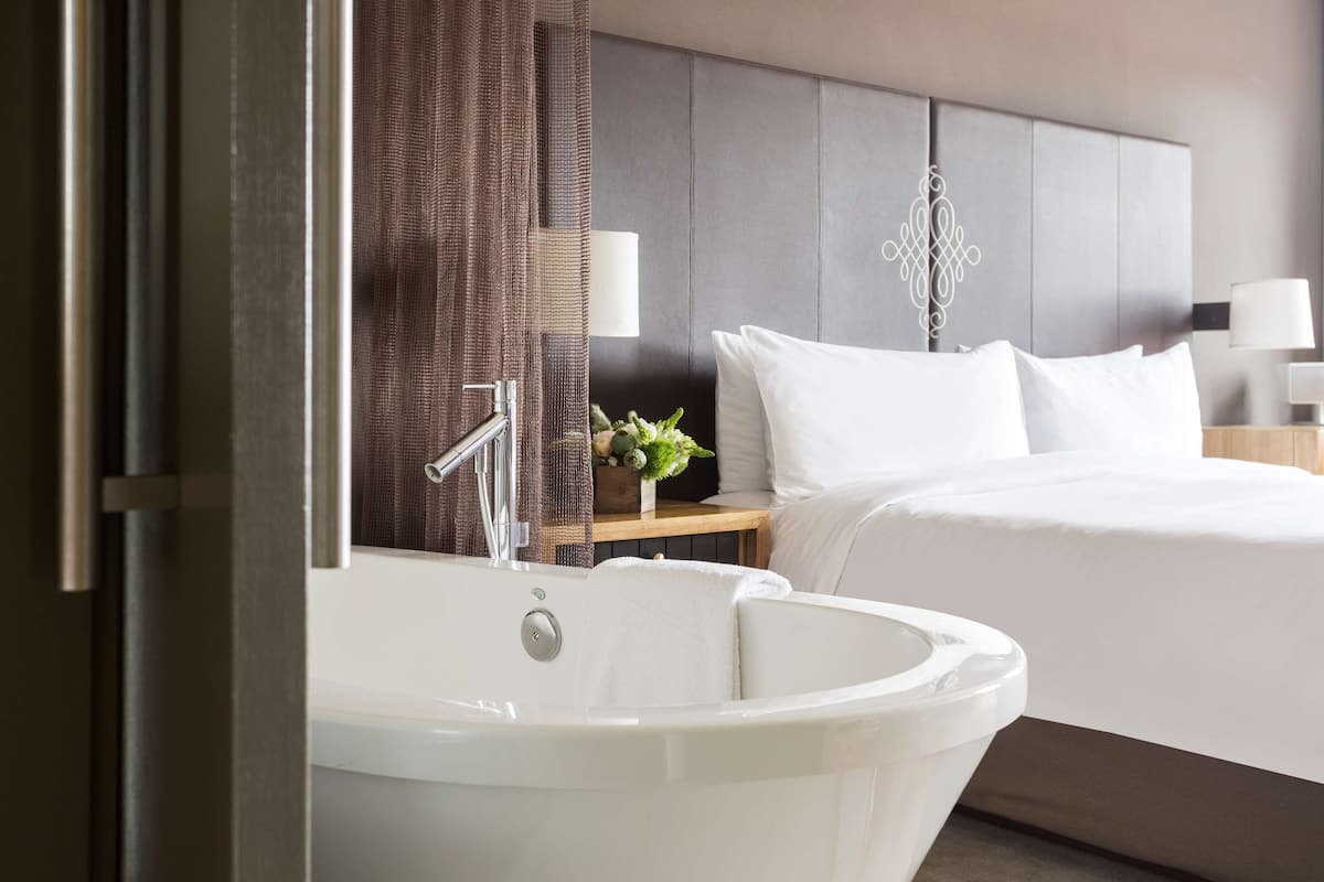 A modern hotel room with a large bathtub, a neatly made bed, and a bedside table with a lamp and a small plant.
