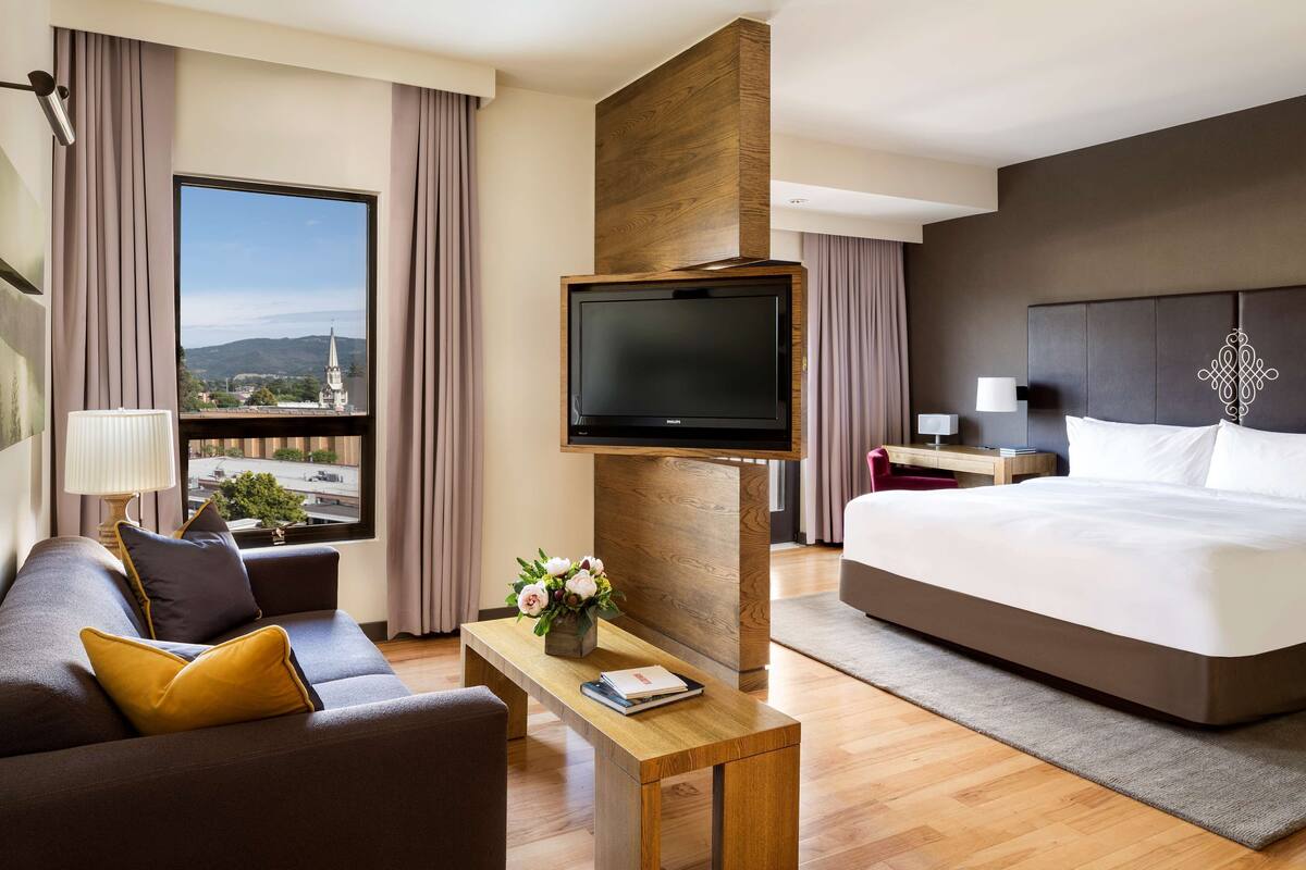 A modern hotel room with a large bed, a sofa, a TV mounted on a wooden panel, a small table with a book and flowers, and a view of a cityscape through large windows.