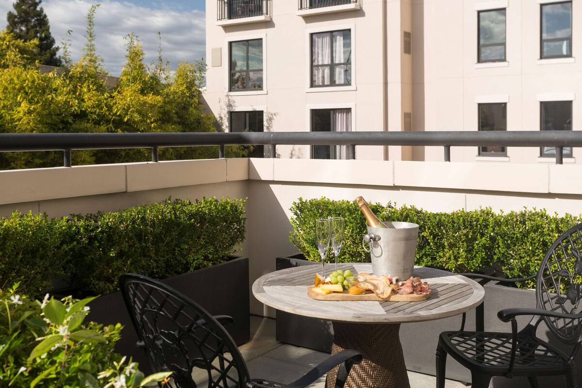 A round table with a bottle of champagne, glasses, and a fruit platter on a balcony with black chairs and a railing.