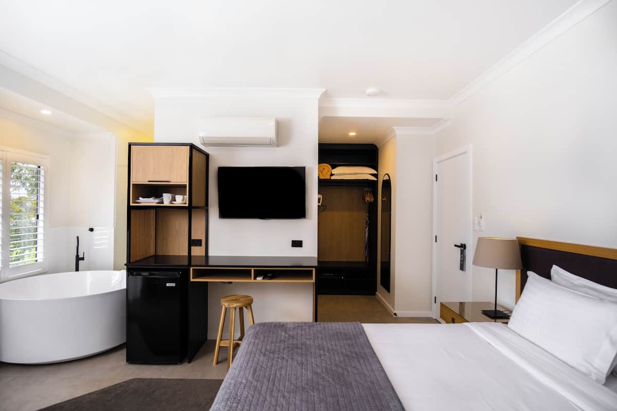 A modern hotel room with a large bed, a desk, a flat-screen TV, a built-in wardrobe, and a separate bathroom with a bathtub.