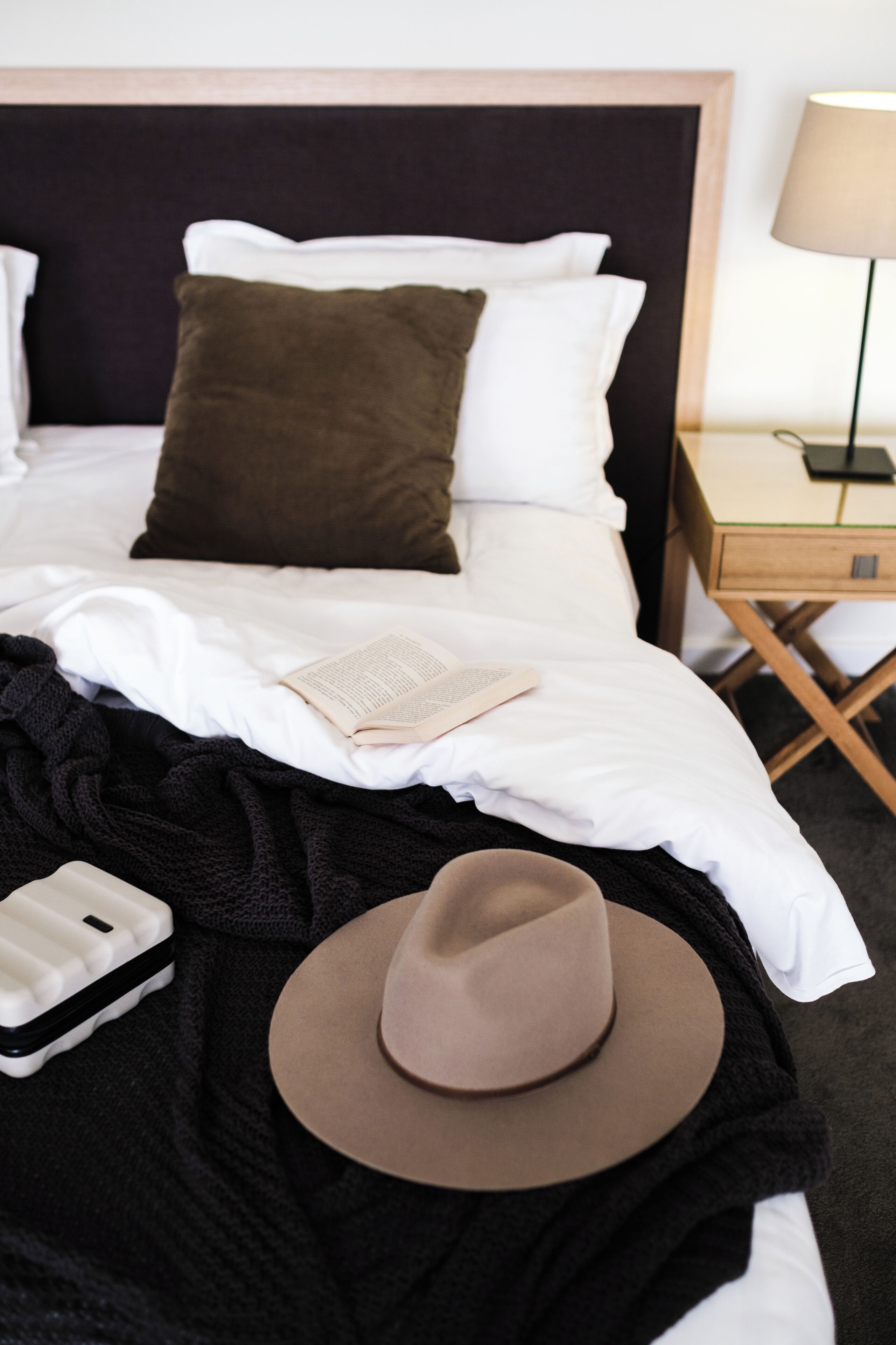 Una cama bien hecha con sábanas blancas, un almohadón marrón, una manta de color marrón oscuro y un sombrero marrón, además de un libro sobre la cama.