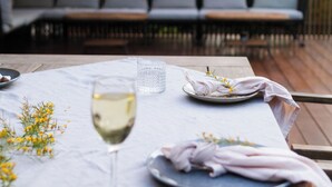 A dining table set with a glass of white wine, a plate with food, and a napkin. There is a bench with cushions in the background.